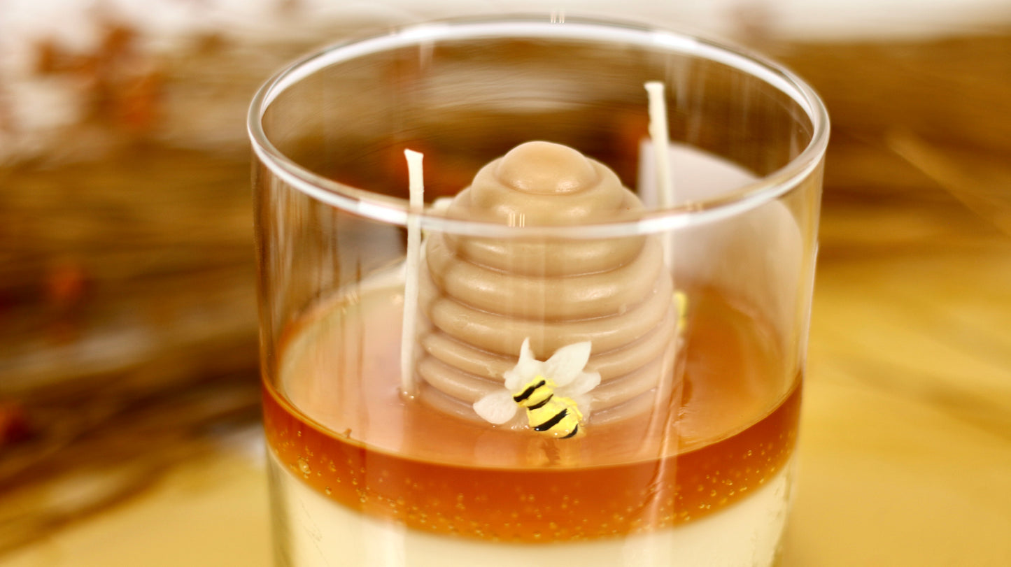 Candle in a glass container with a honeycomb and bee design, set against a blurred natural background.