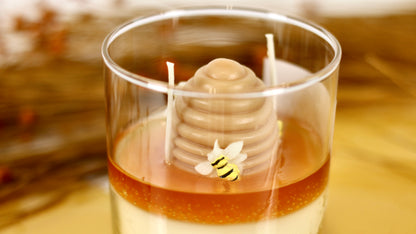 Candle in a glass container with a honeycomb and bee design, set against a blurred natural background.